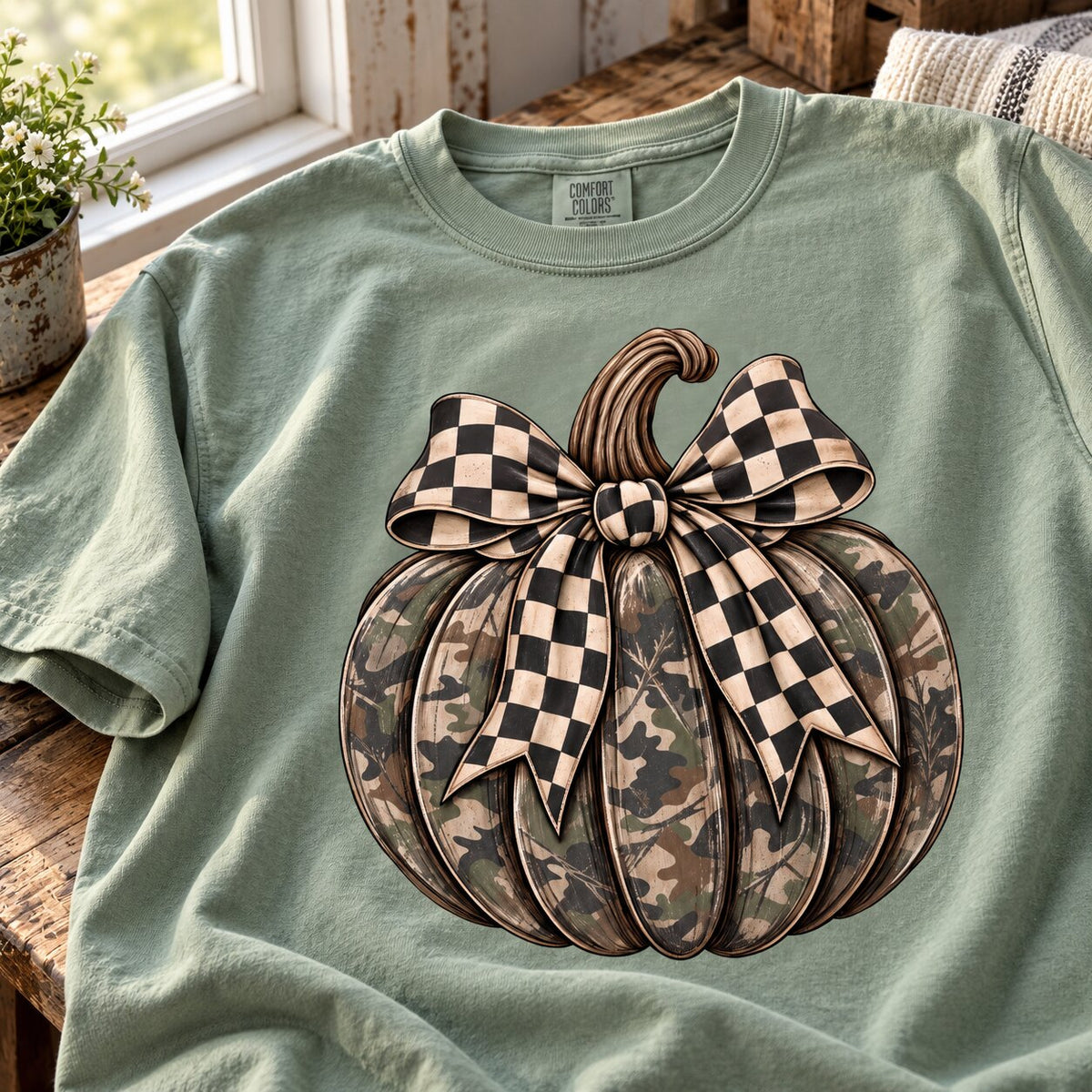 Close-up of the camo pumpkin graphic with a black and cream checkered bow on a sage green tee