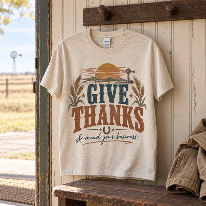 Give Thanks and mind your business tee hanging on a wooden peg on a farmhouse porch wall with a windmill in the background