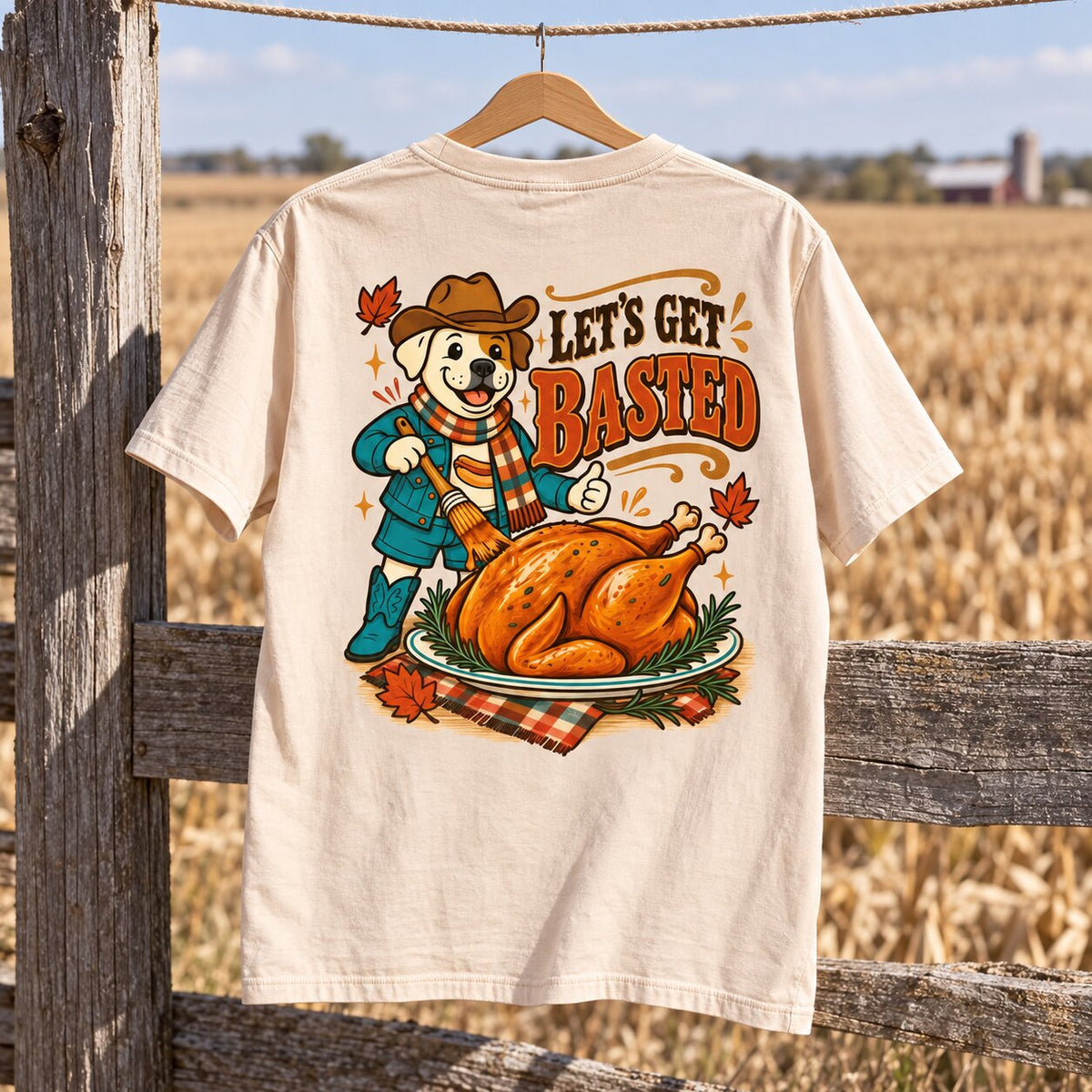 Cream Let's Get Basted tee hanging from a rope line in front of a golden field and weathered fence post