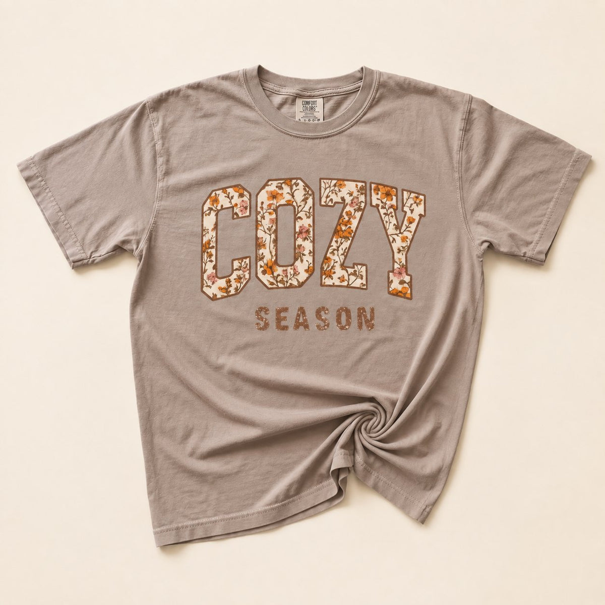 Taupe t-shirt flat lay on a cream background printed with "COZY SEASON" in bold varsity letters filled with an autumn floral pattern.