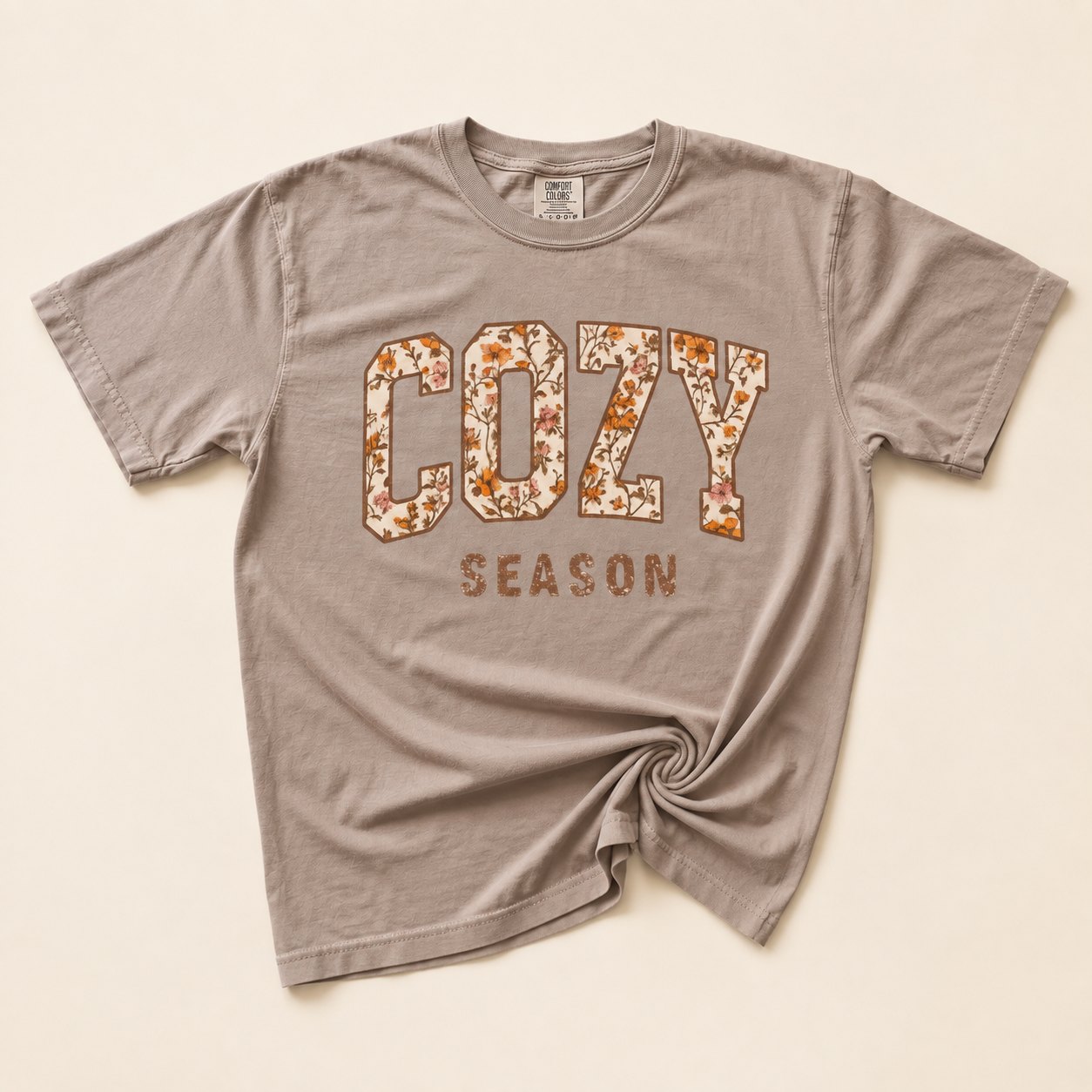 Taupe t-shirt flat lay on a cream background printed with "COZY SEASON" in bold varsity letters filled with an autumn floral pattern.