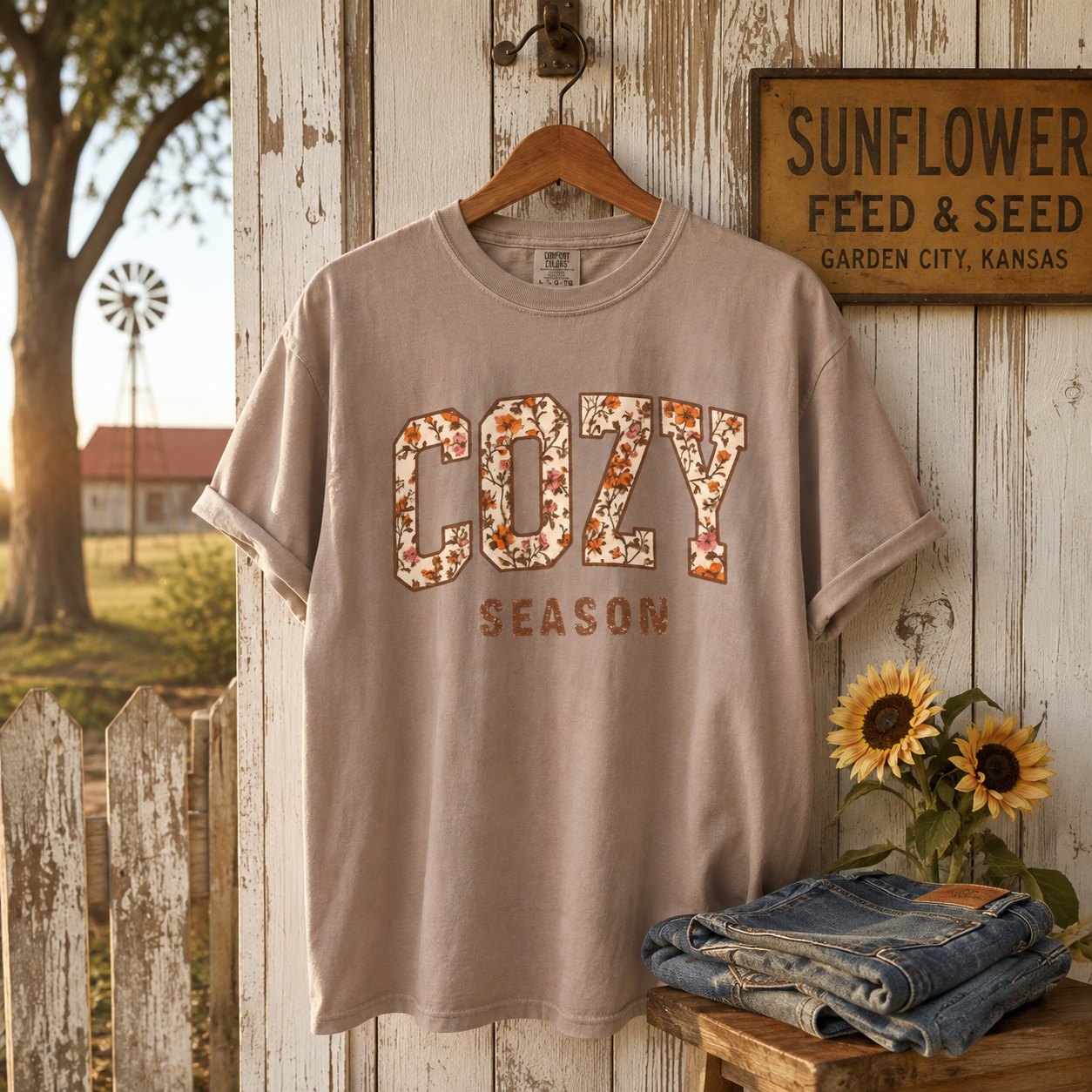 Taupe "Cozy Season" floral varsity t-shirt hanging on a weathered farmhouse door beside a "Sunflower Feed & Seed" sign, sunflowers, and folded denim.