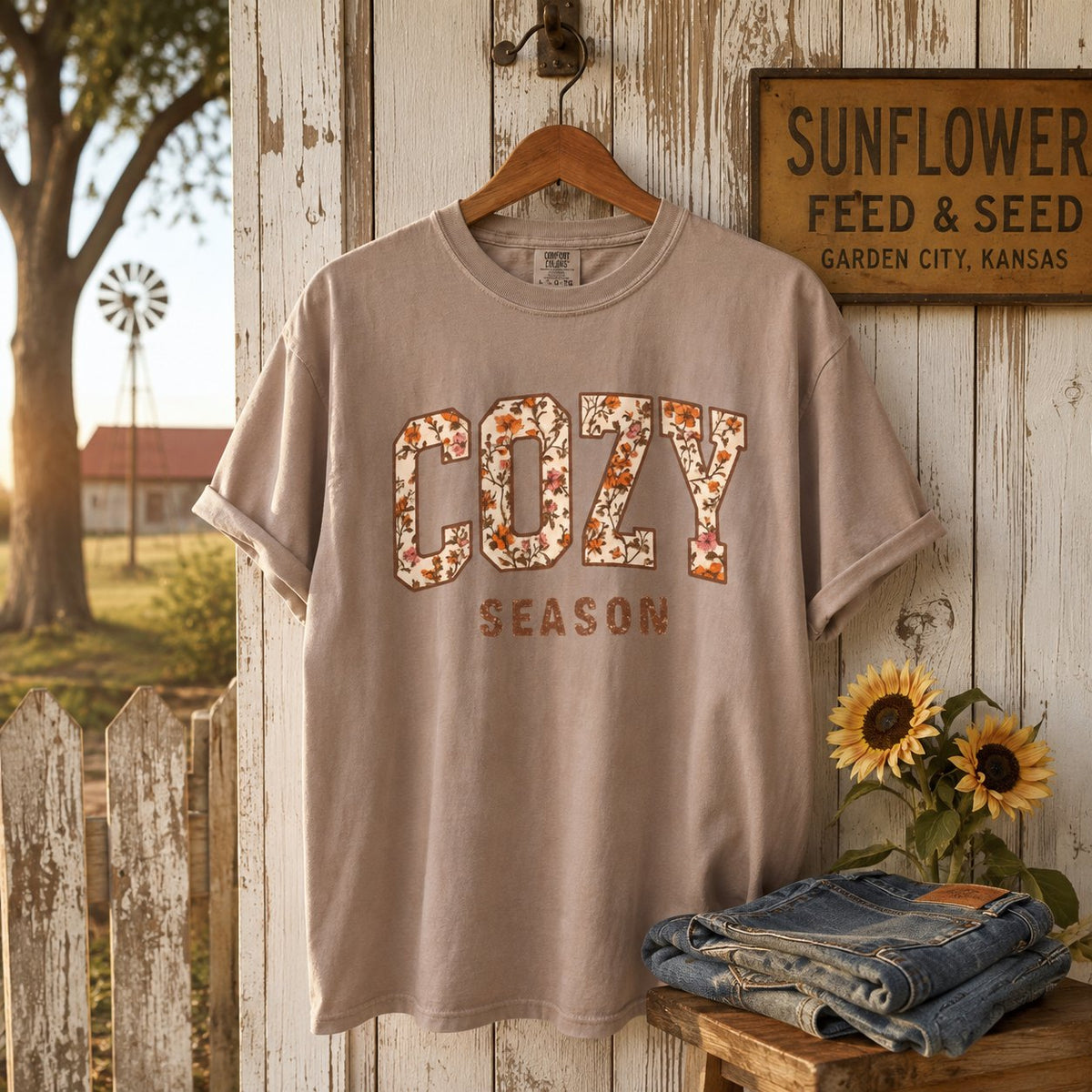 Taupe "Cozy Season" floral varsity t-shirt hanging on a weathered farmhouse door beside a "Sunflower Feed & Seed" sign, sunflowers, and folded denim.