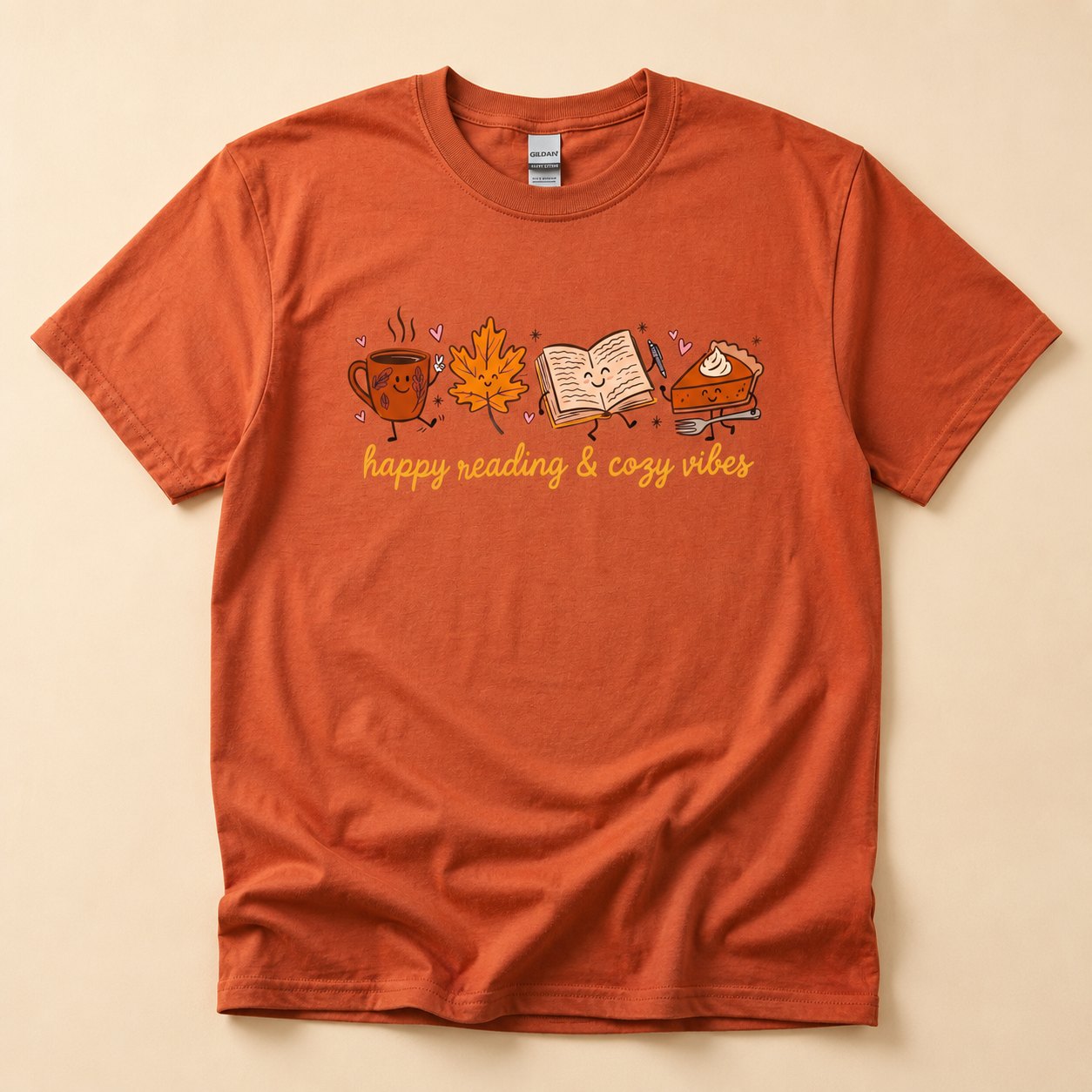 Burnt orange t-shirt flat lay on a cream background printed with cartoon characters of a coffee mug, maple leaf, book, and pumpkin pie slice above the text "happy reading & cozy vibes."