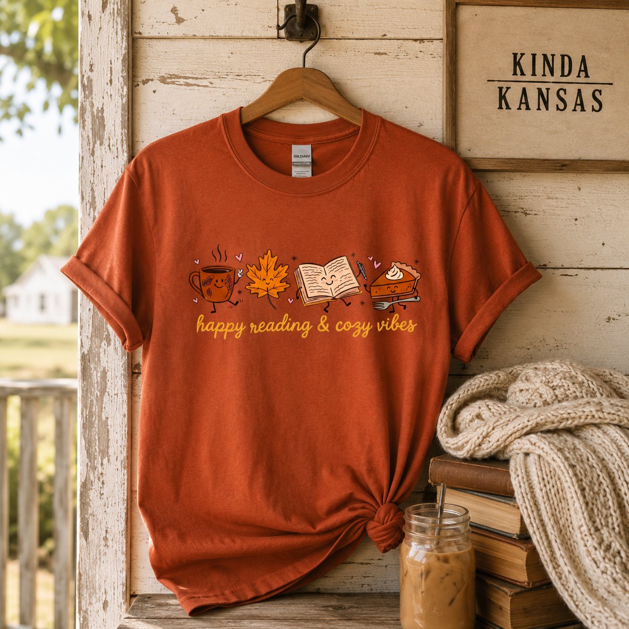 Burnt orange "happy reading & cozy vibes" t-shirt hanging on a rustic porch hook beside a "Kinda Kansas" sign, a stack of books, an iced coffee, and a knit blanket.