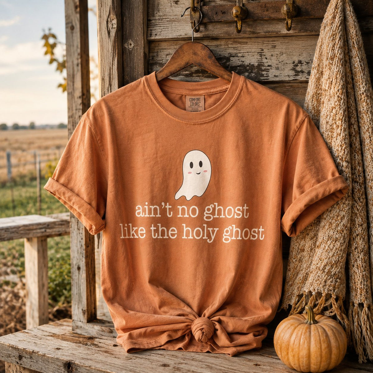 Rust orange "ain't no ghost like the holy ghost" t-shirt hanging on a rustic porch hook beside a knit throw and a small pumpkin.