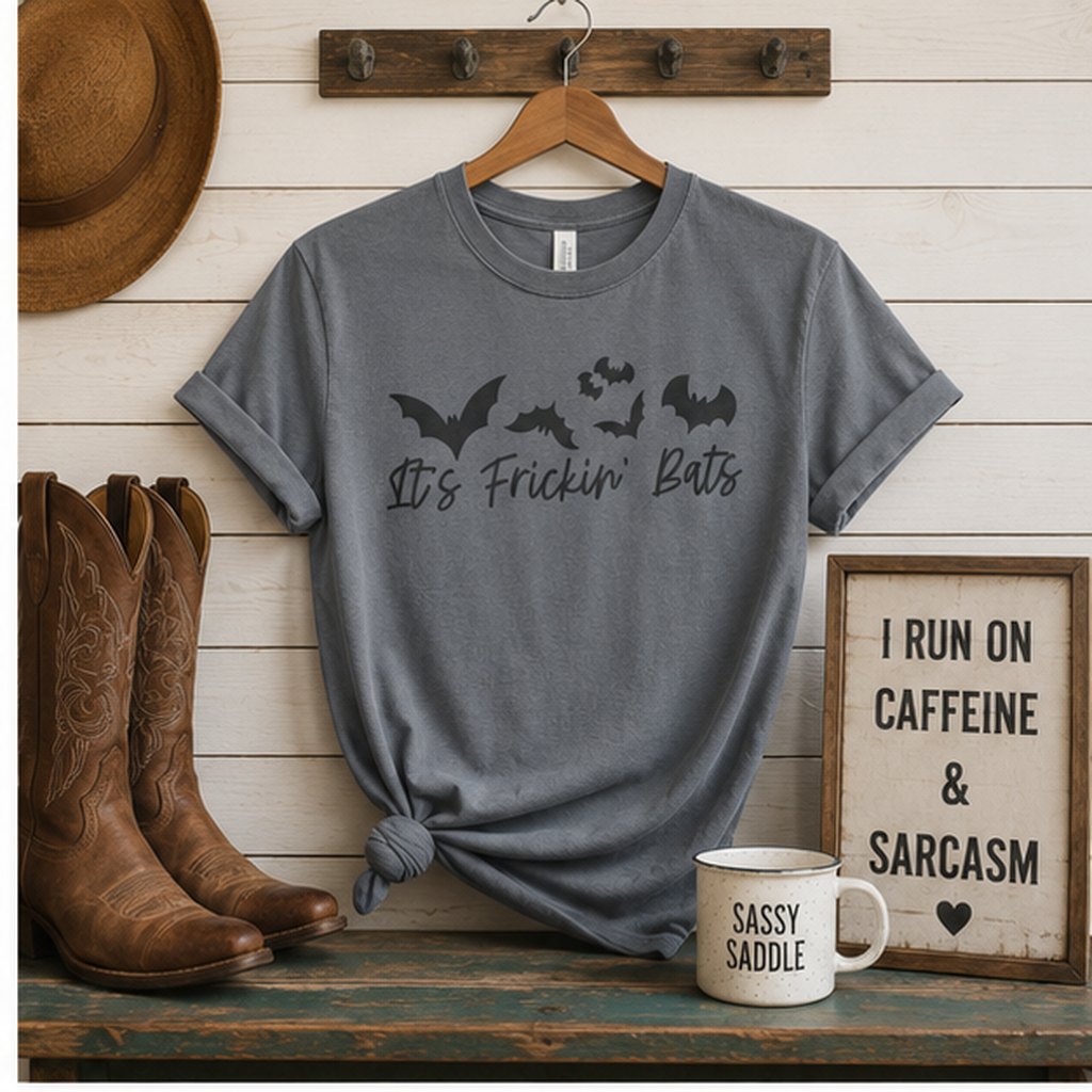 Charcoal grey "It's Frickin' Bats" t-shirt hanging on a wooden coat hook above cowboy boots, a straw hat, and a Sassy Saddle mug on a rustic farmhouse shelf.