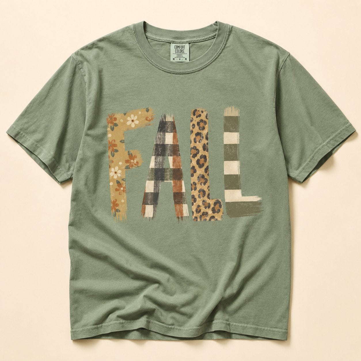 Sage green t-shirt flat lay on a cream background printed with the word "FALL" in mixed patchwork textures: floral, buffalo check, leopard print, and stripes.