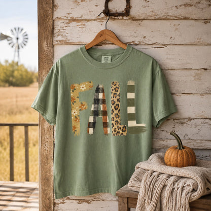 Sage green patchwork "FALL" t-shirt hanging on a weathered farmhouse porch post beside a knit blanket and a small pumpkin.