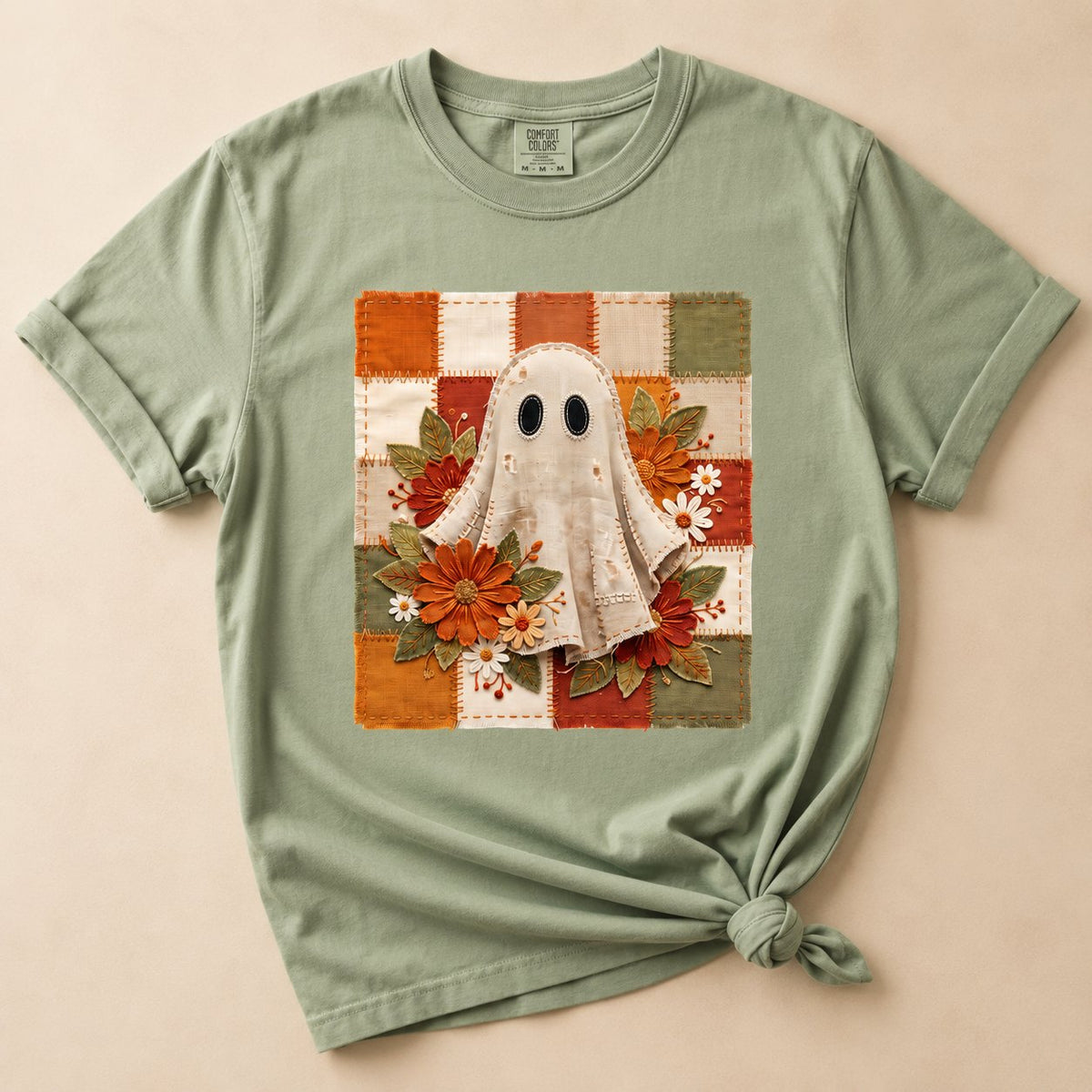Sage green t-shirt flat lay on a cream background printed with a stitched-fabric ghost surrounded by orange and rust fall florals on a patchwork checkerboard square.