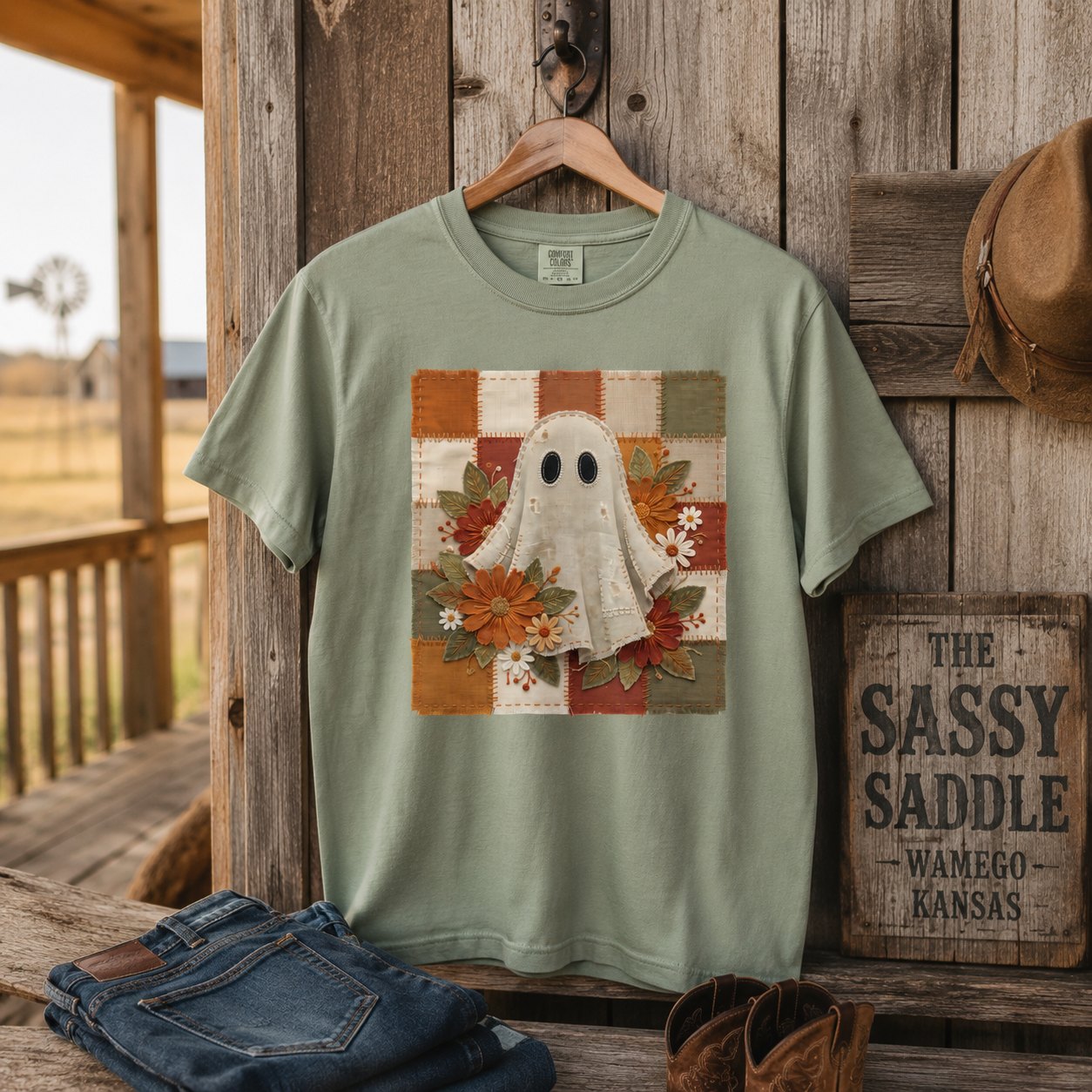 Sage green patchwork ghost t-shirt hanging on a rustic wooden porch beside a "The Sassy Saddle, Wamego Kansas" sign, cowboy boots, and folded denim.