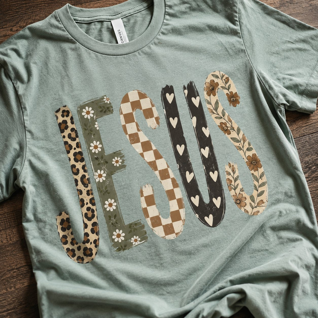 Close-up of the patchwork "JESUS" graphic showing the leopard, daisy, checkerboard, heart, and floral letter textures on sage green cotton.