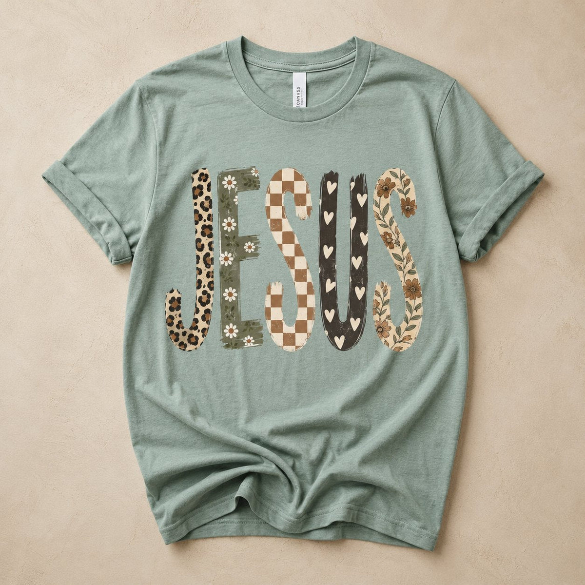 Sage green t-shirt flat lay on a tan background printed with the word "JESUS" in mixed patchwork textures: leopard print, daisy floral, checkerboard, hearts, and vine florals.