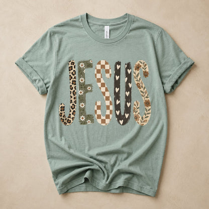 Sage green t-shirt flat lay on a tan background printed with the word "JESUS" in mixed patchwork textures: leopard print, daisy floral, checkerboard, hearts, and vine florals.