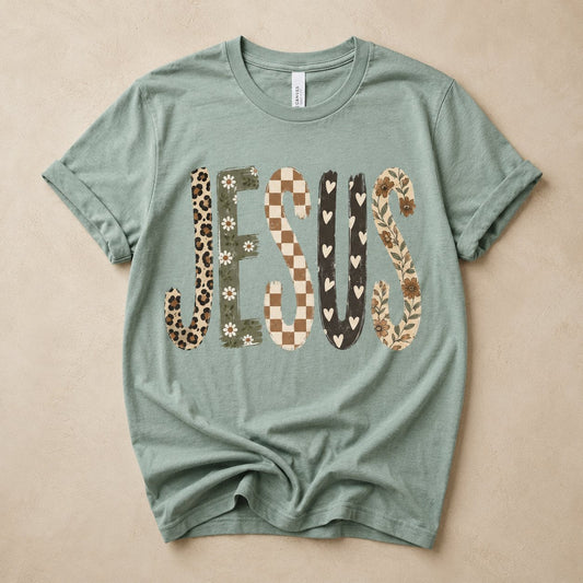 Sage green t-shirt flat lay on a tan background printed with the word "JESUS" in mixed patchwork textures: leopard print, daisy floral, checkerboard, hearts, and vine florals.