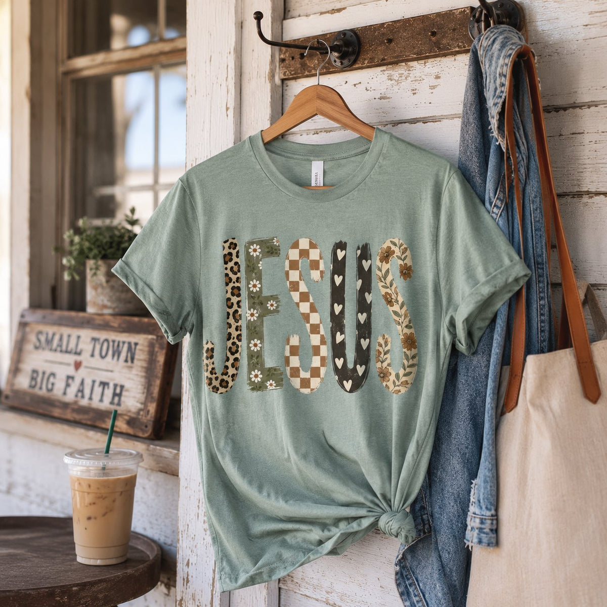 Sage green patchwork "JESUS" t-shirt hanging on a rustic porch hook beside a "Small Town Big Faith" sign, an iced coffee, and a canvas tote.