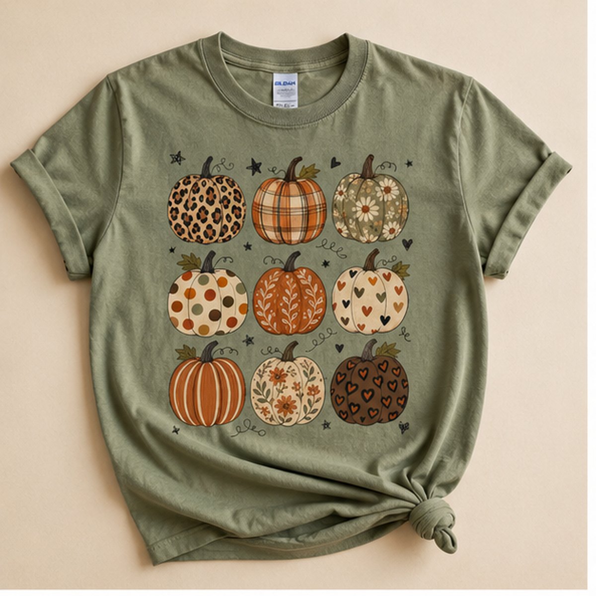 Sage green t-shirt knotted at the hem, flat lay on a cream background, printed with a 3x3 grid of nine differently patterned pumpkins (leopard, plaid, daisy, polka dot, floral, striped, and heart print).