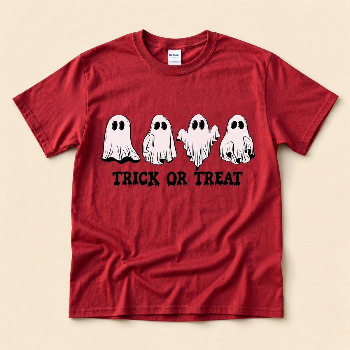 Red t-shirt flat lay on a cream background printed with four hand-drawn ghosts in different poses above dripping "TRICK OR TREAT" lettering.