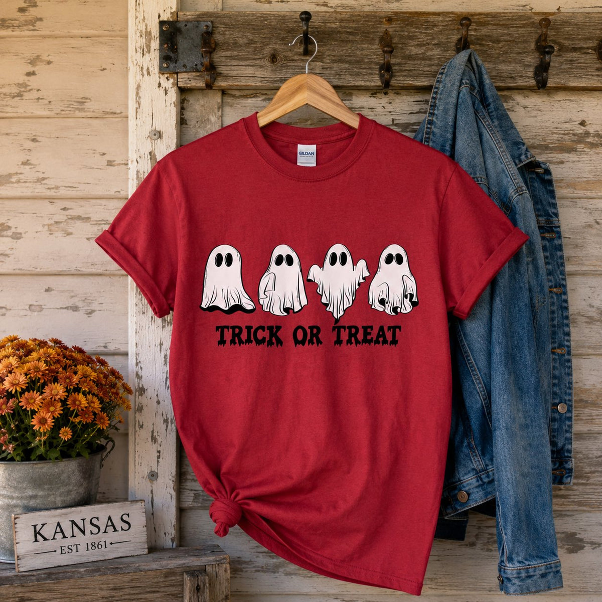 Red "Trick or Treat" ghost t-shirt hanging on a rustic door beside a denim jacket, mums, and a "Kansas Est. 1861" sign.