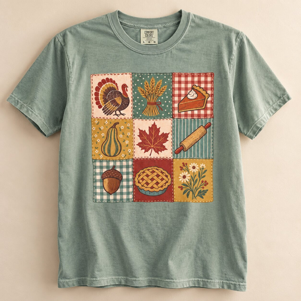 Sage green Thanksgiving patchwork graphic tee laid flat, with a quilt-style grid of turkey, wheat, pie, maple leaf, rolling pin, acorn and wildflower squares